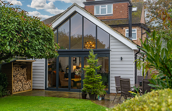 external view of addition to a famliy home with floor to eaves glazing and pale blue/grey cladding nestled into a vibrant green garden
