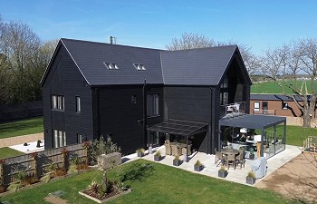 Image of Whinhams farm - newly extended property with clean lines, in a modern agricultural style with sleek black cladding. The building is set in newly landscaped gardens against a blue sky and the fields beyond.